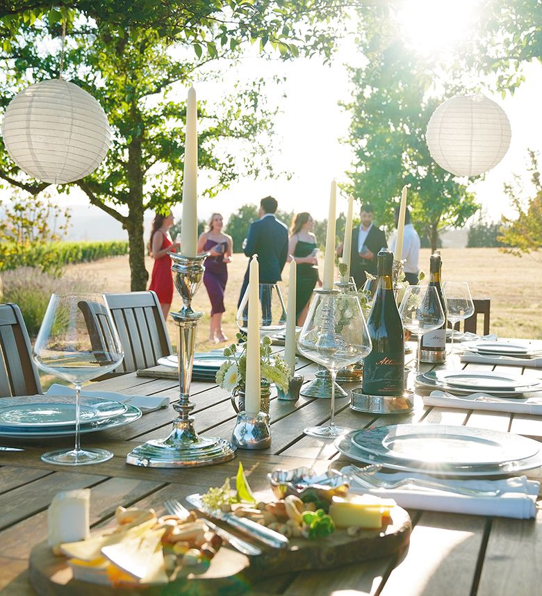 Placesettings and wine glasses on a table while a group of people stand nearby.  ## Photo by IFM Photography