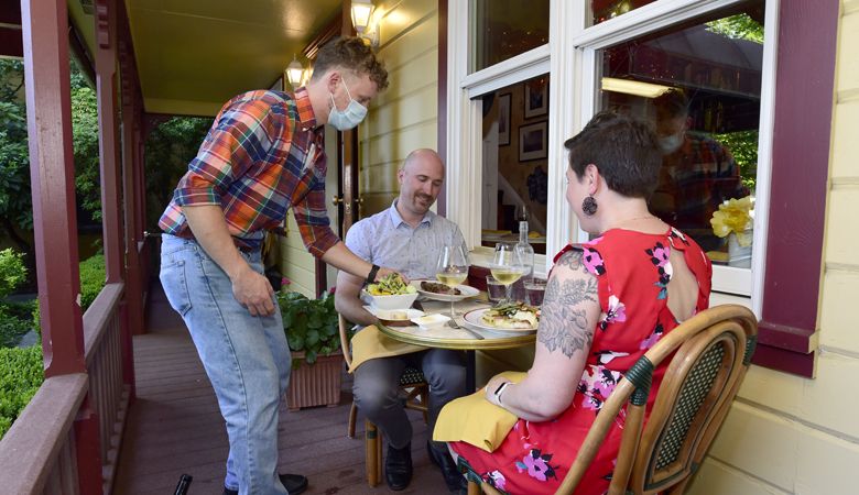 A masked server at Bistro Maison in McMinnville delivers entrées to a couple sitting on the elegant porch located around the corner from the patio. ##Photo by Marcus Larson