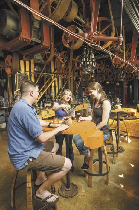 A Quenett staff member pours glasses of wine for a couple visiting the winery’s tasting room located inside the Sunshine Mill in The Dalles. Pulls and levers, leftover from the Mill’s former life as a flour mill, create an industrial chic vibe throughout the entire structure. Photo by Andrea Johnson.