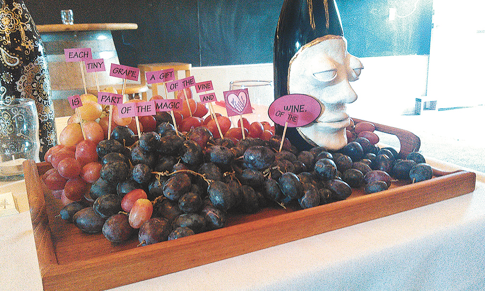 Two artist-decorated bottles stand guard over a tray of tasty grapes at the Drabkin auction. Sequential signs on toothpicks read “Each tiny grape is a gift of the vine and a part of the magic of the wine.” ##Photo provided